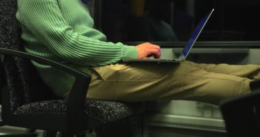 Loş tren vagonunda dizüstü bilgisayar, yeşil süveter ve haki pantolon, klavyede eller, pencere.