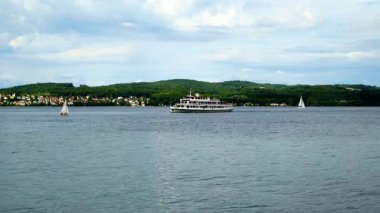 Büyük bir feribot gölde ilerlerken yelkenli tekneler suda süzülüyor. Yeşil tepeler arka planda gri bulutların altında yükseliyor. Suda yoğun bir gün..