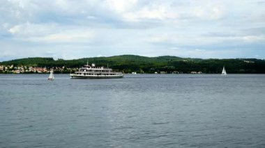 Küçük yelkenliler süzülürken büyük bir tekne gölün karşısına geçiyor. Arkaplanda bulutlu bir gökyüzünün altında yeşil tepeler yükseliyor. İnsanlar su kenarında vakit geçirmeyi sever..