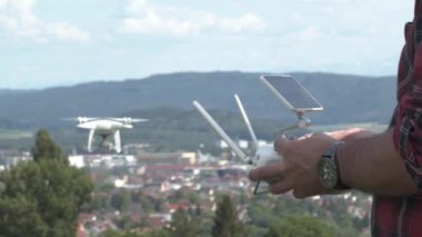 Bir insan bir tepede duruyor ve uzaktan kumandalı bir dron tutuyor. İnsansız hava aracı, aşağıdaki bir şehrin üzerinde, tepelerle birlikte havada süzülüyor. Hava açık ve gökyüzü parlak..