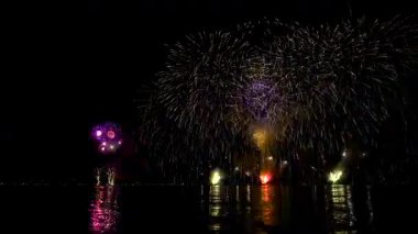 Parlak havai fişekler gölün üzerinde gece gökyüzünü aydınlatıyor. Renkler patlıyor ve sakin suya yansıyor. İnsanlar sahilde izleyip kutluyor..