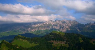 Altında karla kaplı tepeler ve yeşil vadilerle güzel bir dağ manzarası. Bulutlar arazide sürükleniyor, dinamik bir gökyüzü yaratıyor. Doğa bu alandaki çeşitliliğini gösteriyor..