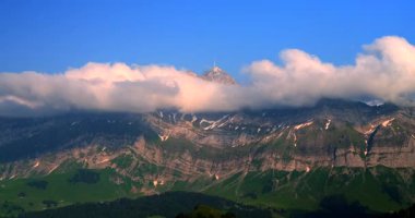 Zirvesinde sürüklenen bulutlarla bir dağ dimdik ayakta duruyor. Güneş kayalık yüzeyde parlıyor, hava açık ve parlak kaldıkça aşağıdaki yeşil araziyi aydınlatıyor..