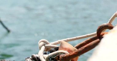 Rıhtımın yanında su yavaşça akar. Bir ip parçası paslı bir zincire bağlı. Güneş suyun üzerinde parlıyor, yansımalar yaratıyor. Tekneler uzaktan görülebilir..