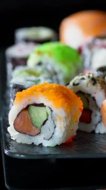 Suşi ruloları siyah bir tabakta düzenlenir. Tabakta çeşitli renkler ve malzemelerle karışık suşi türleri yer alıyor. Yoğun bir restoran ortamında insanlar yemekleri severler..