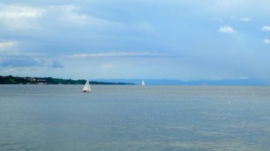 Kalın bulutlar gökyüzünü kaplarken iki yelkenli denizde yüzüyor. Göl kenti uzaktan görülebiliyor ve akşam saatlerinde manzaralı bir manzara yaratıyor..