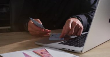 Bir kişi elinde dizüstü bilgisayarla bir masada otururken nakit paraları sıralıyor. Öğleden sonra doğal ışık alan bir ev ofisi var..