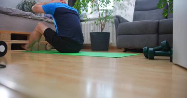 Bir adam oturma odasında yeşil bir paspasın üzerinde oturuyor. Sabah rutininin bir parçası olarak egzersiz yapıyor. Odada bitkiler, ağırlıklar ve rahat bir sandalye var..