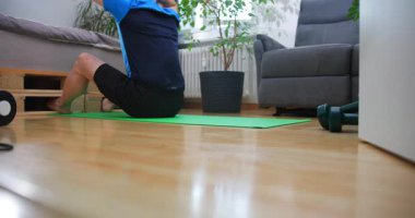 Bir insan oturma odasındaki yeşil yoga minderinin üzerinde egzersiz yapıyor ve esniyor. Yerdeki ağırlıklar görülebilirken yakınlarda bitkiler ve bir kanepe var..