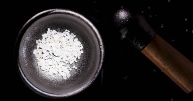 Tahta bir tokmak yakınlarda dinlenirken bir kase ezilmiş tuz içerir. Dekor, akşam yemeği oturumunu çağrıştıran karanlık bir yüzeyi gösteriyor. Sahne, basit mutfak aktivitelerini vurguluyor.