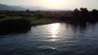 Güneş batarken, onun ışınları yoğun Akdeniz ormanlarında parlıyor ve sakin denize dokunuyor. Sakin dalgalar Yunanistan 'daki bu sakin kıyı sahnesindeki ışığı yansıtıyor.