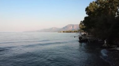 Taşları ve sakin suları olan ücra bir kumsal yoğun Akdeniz bitkilerinin gölgesinde uzanıyor. Ağaçlar yumuşak dalgaların üzerine uzanırken kuru dallar berrak sularda dinleniyor. Uzakta, dağlar bu huzurlu kıyı manzarasını oluşturur..