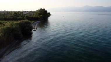 Küçük bir köyün yakınında uzun ağaçlar ve yoğun Akdeniz bitkileri arasında gizli bir sahil bulunur. Sakin mavi deniz, güneş Yunanistan 'ın uzak dağlarının ardında batarken ışığı yansıtıyor.