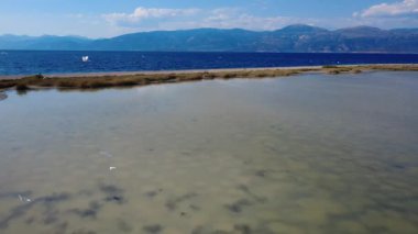 Kuşlar Akdeniz yakınlarında kayalık tepelerle çevrili bir lagünün üzerinde süzülür. Sakin su, Yunanistan 'ın mavi gökyüzünü yansıtıyor