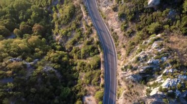 Yunanistan 'ın dağlık kesimindeki izole edilmiş asfalt ulusal karayolunun dikey hava aracı görüntüsü, günbatımında dik kayalık yamaçlardaki sık ormanı geçiyor. Trafik yok. Beyaz çizgiler geçiş izni olmadığını gösteriyor. Uçurumun kenarındaki koruyucu güvenlik bariyerleri.