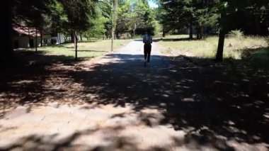 Genç bir kadın gölgeli bir şehir parkında uzun ağaçlarla yavaşça yürüyor. Dağlık alana bakarken birini bekliyor..