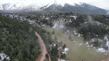 Yüksek görüş açısı, park halindeki araçlar, yoğun köknar ormanı, karlı bölgeler ve Yunanistan, Avrupa 'daki engin bir vadinin üzerindeki uzak tepelerin bulunduğu sisli bir dağ tarlasında serpentin toprak bir yol sunuyor..