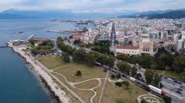 Yunanistan ve Avrupa 'da mavi gökyüzü ve dağ zeminine karşı deniz kenarı gezinti alanı, liman tesisleri ve merkez kilise kulesini ön plana çıkaran geniş şehir manzarası.