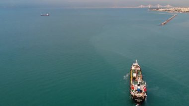 Küçük ticari tanker, görünür limanı ve dalgakıranı olan endüstriyel liman şehrine yaklaşırken durgun turkuaz sularda uyanır..