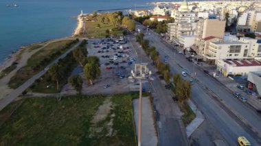 Yeşil parkı ve otoparkı olan binaların yanındaki şehir sahil bulvarında. Liman altyapısı ve sakin deniz arka planda görünüyor..