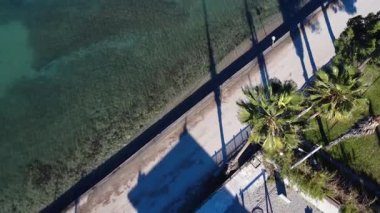 Palmiye ağaçları, deniz kenarındaki turkuaz turkuaz Akdeniz suyunun yanında kum taşları ve deniz yosunlarıyla kaplı bir yol üzerindedir. Yunanistan ve Avrupa 'da yaz ortasında hafif dalgalar yüzey boyunca hareket ediyor.