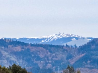 Sonbaharın sonları ile kışın başlangıcı arasındaki zıtlığı gösteren bir dağ manzarası. Ön planda ve orta kısımda mavi, yoğun ormanlık tepeler bulunur. Yaprakların koyu mavi tonları havadan bir perspektif yaratıyor. İçeri