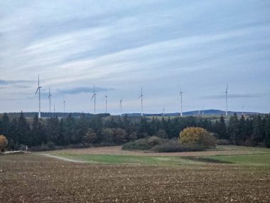 Gün batımında ya da alacakaranlıkta çekilen bu fotoğraf ufukta yükselen uzun rüzgar türbinlerini gösteriyor. Rüzgar jeneratörleri uzak bir tepenin ya da yaylanın üzerinde yer alıyor. Bu yaylalar yoğun, koyu yeşil bir orman şeridi tarafından kesiliyor.