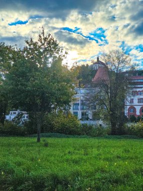 Kentsel mimarinin yenilenmiş sanayi tarzı binalar ve modern daireleri birleştirdiği manzara. Önde yeşil çim ve nehir, arkada orman tepesi..