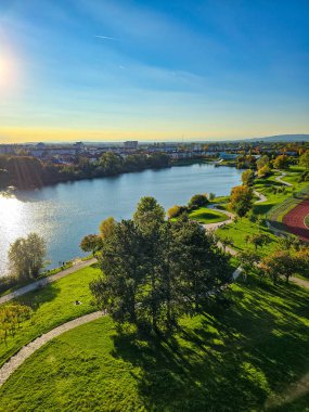 Sonbahar boyunca kentsel binalar arasında renkli turuncu ve sarı yaprakların yer aldığı manzaralı bir şehir. Arka planda, orman tepeleri ve rüzgar türbinleri dağınık bulutlarla mavi bir gökyüzünün altında görülebilir..