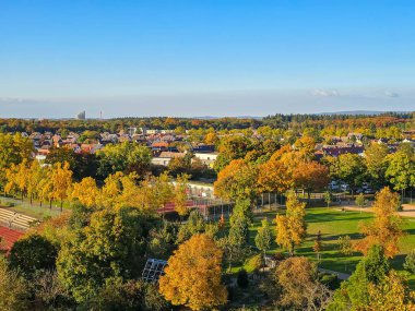Sonbahar boyunca kentsel binalar arasında renkli turuncu ve sarı yaprakların yer aldığı manzaralı bir şehir. Arka planda, orman tepeleri ve rüzgar türbinleri dağınık bulutlarla mavi bir gökyüzünün altında görülebilir..