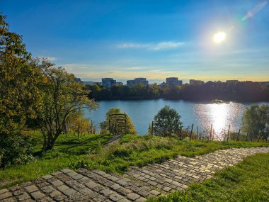 Sonbahar boyunca kentsel binalar arasında renkli turuncu ve sarı yaprakların yer aldığı manzaralı bir şehir. Arka planda, orman tepeleri ve rüzgar türbinleri dağınık bulutlarla mavi bir gökyüzünün altında görülebilir..