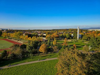 Sonbahar boyunca kentsel binalar arasında renkli turuncu ve sarı yaprakların yer aldığı manzaralı bir şehir. Arka planda, orman tepeleri ve rüzgar türbinleri dağınık bulutlarla mavi bir gökyüzünün altında görülebilir..