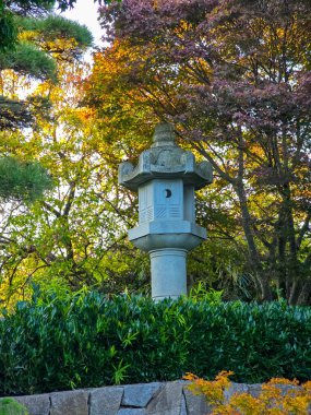 Zarif, uzun granit bir fener canlı sonbahar yaprakları ve zen bahçesindeki yeşil çalıların arasında duruyor. Sıcak akşam ışığı taşın karmaşık oymalarını ve çevreleyen doğanın zengin renklerini güçlendirir..