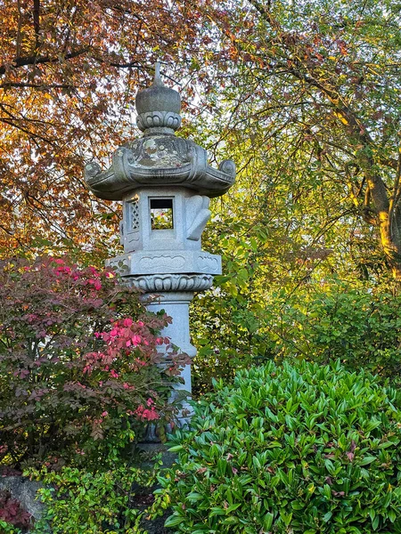 Zarif, uzun granit bir fener canlı sonbahar yaprakları ve zen bahçesindeki yeşil çalıların arasında duruyor. Sıcak akşam ışığı taşın karmaşık oymalarını ve çevreleyen doğanın zengin renklerini güçlendirir..