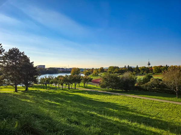 Güneşli bir günde canlı yeşil bir parkın geniş açılı manzarası. Sahnede yuvarlanan çimenli tepeler, asfalt bir yürüyüş yolu, dingin bir göl ve ufukta mavi bir gökyüzünün altında yumuşak ışık ışınlarıyla yüksek bir iletişim kulesi yer alıyor..