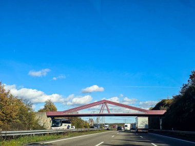 Aydınlık bir günde çok şeritli otobanın geniş bir görüntüsü. Birkaç kamyon ve yolcu vagonu asfalt yolda ilerliyor. Yol kenarında sonbahar ağaçları ve beton bir güvenlik bariyeri var. Yüksek voltajlı elektrik hatları görünür.