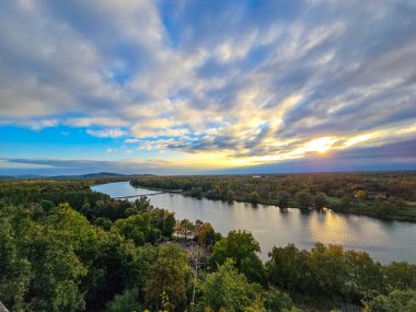 Sonbahar mevsiminde gür bir ormanın içinden geçen geniş bir nehrin yüksek açılı manzarası. Güneş canlı bulutların ardında batıyor, suda altın bir yansıma yaratıyor ve huzurlu, sakin bir atmosfer yaratıyor..