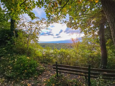 Orman yamacına kurulmuş ahşap bir park bankının olduğu huzurlu yüksek açılı bir manzara. Sahne, canlı yeşil ve sarı akçaağaç yapraklarıyla güzel çerçevelenmiş, yumuşak bir günbatımı gökyüzünün altındaki geniş bir nehir vadisine bakıyor. Her yer sonbahar kaplandı.