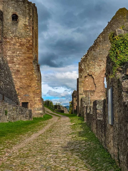 Hochburg Kalesi 'nin yüksek ortaçağ duvarlarının geniş açılı görüntüsü. Geleneksel bir kaldırım taşı yolu, Kara Orman bölgesinde dramatik bir gökyüzüne karşı inşa edilen muazzam tahkimatların yanında yer alıyor..