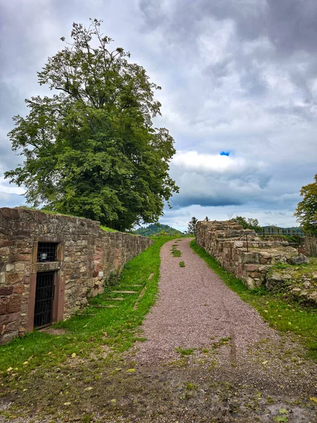 Hochburg Kalesi 'nin yüksek ortaçağ duvarlarının geniş açılı görüntüsü. Geleneksel bir kaldırım taşı yolu, Kara Orman bölgesinde dramatik bir gökyüzüne karşı inşa edilen muazzam tahkimatların yanında yer alıyor..