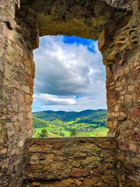 Hochburg şatosu harabelerinin kalın savunma duvarında açılan engebeli bir taş pencerenin yakın çekim görüntüsü. Küçük yabani bitkiler bulutlu bir gökyüzüne karşı antik duvarcılıktan büyürler..