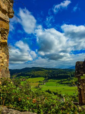 Ortaçağ kalesinin taş duvarlarından manzara manzarası. Canlı yeşil bir vadi, küçük bir köy, tarım arazileri, ve dramatik mavi gökyüzünün altında yuvarlanan tepeler yaz veya sonbaharda beyaz bulutlar.