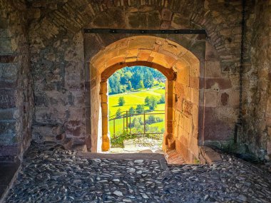 Ortaçağ şatosu ya da kalesinin antik taş kemerinden bir manzara, parlak güneş ışığı altındaki tarlaları ve ormanları olan canlı yeşil bir vadiye götürüyor. Ön planda kaldırım taşı zemin ve dokulu kumtaşı duvarlar var, doğal bir çerçeve oluşturuyor.