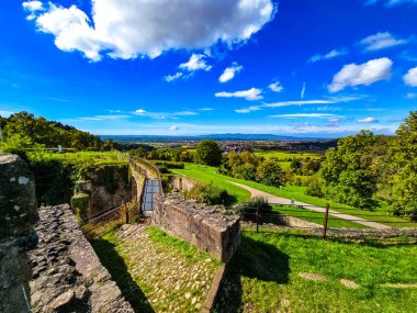 Yıpranmış duvarları ve yeşil bitki örtüsüyle antik taş kalenin manzarası, Avrupa 'da parlak bulutlu bir gökyüzü altında kayan tepeler ve dağlardan oluşan panoramik bir manzaraya ev sahipliği yapıyor..
