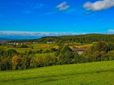 Canlı yeşil bir vadiye yerleşmiş manzaralı kırsal bir köyün yüksek açılı manzarası. Ön planda, dağınık ağaçlarla kaplı yuvarlanan çimenli tepeler gözü yerleşim yerine yönlendirir ve görkemli dağ geniş mavi ve beyaz bulutlu gökyüzünün altında sıralanır. Bu...