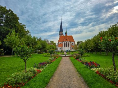 Manikürlü bir parktaki yüksek kuleli tarihi Gotik tarzı bir kilisenin manzarası. Ön planda canlı mor ve sarı çiçekler yer alırken, yemyeşil çimenler ve sıra sıra ağaçlar gözü mimariye yönlendiriyor.