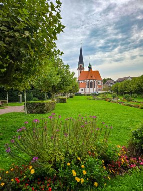 Manikürlü bir parktaki yüksek kuleli tarihi Gotik tarzı bir kilisenin manzarası. Ön planda canlı mor ve sarı çiçekler yer alırken, yemyeşil çimenler ve sıra sıra ağaçlar gözü mimariye yönlendiriyor.