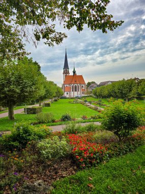 Manikürlü bir parktaki yüksek kuleli tarihi Gotik tarzı bir kilisenin manzarası. Ön planda canlı mor ve sarı çiçekler yer alırken, yemyeşil çimenler ve sıra sıra ağaçlar gözü mimariye yönlendiriyor.