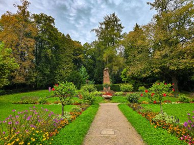 Yeşil ağaçların arasına yerleşmiş geleneksel ahşap bir çardağın yer aldığı Avrupa parkının güzel bir manzarası. Ön planda düzgün budanmış çalılar, küresel çalılar ve bahçe yolları bulutlu bir günde huzurlu ve sakin bir atmosfer yaratıyor..