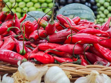 Ön planda canlı ve renkli kırmızı biber yığını. Altında da sarımsak başları var. Arka planda, çeşitli koyu yeşil sebzeler, zengin ve iştah açıcı bir kompozisyon yaratarak yemek pişirme ve şamata yapma sevincini uyandırıyor.